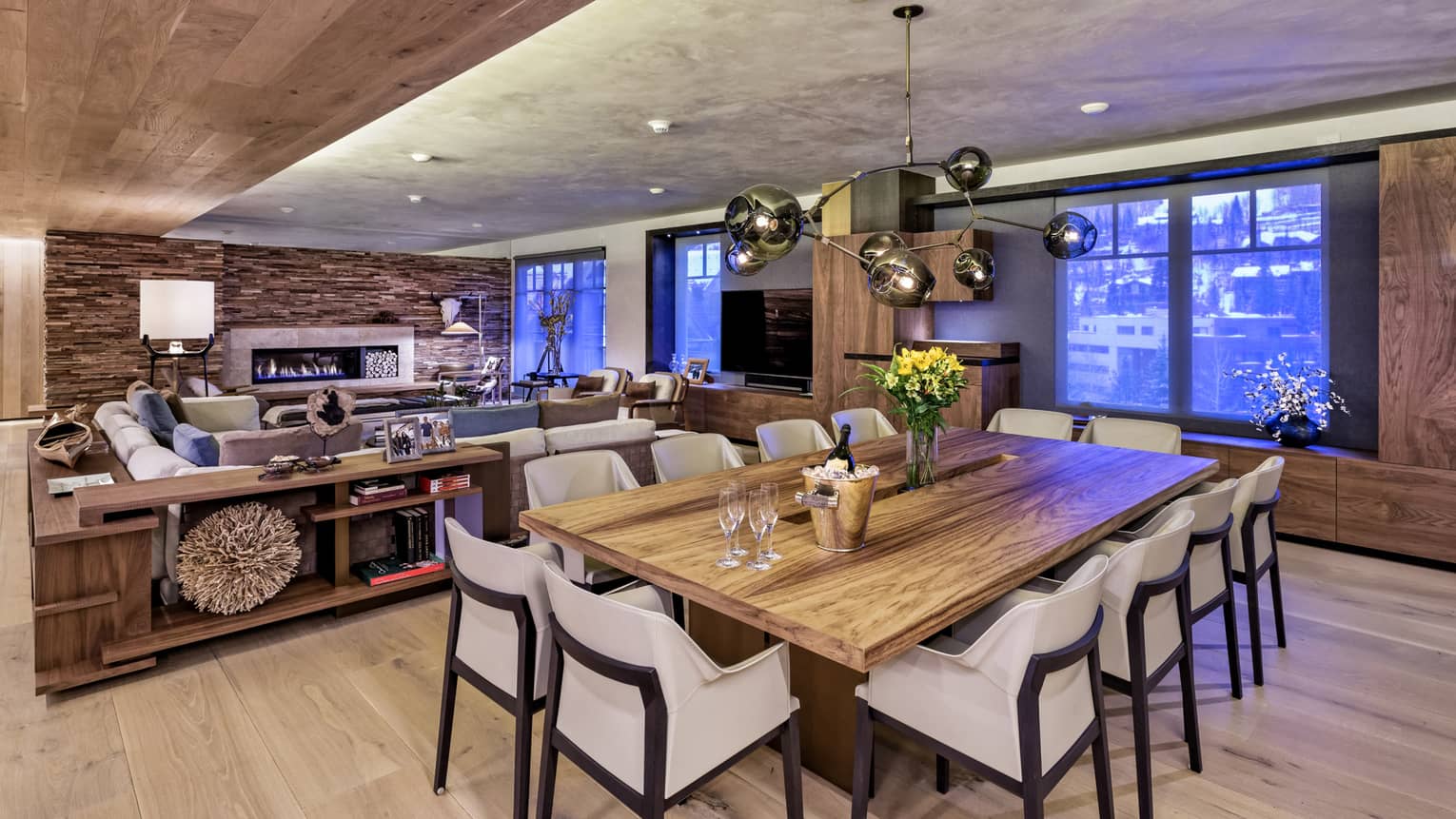 Modern wood farm-style dining table lined with white chairs, Champagne on ice, in front of living room