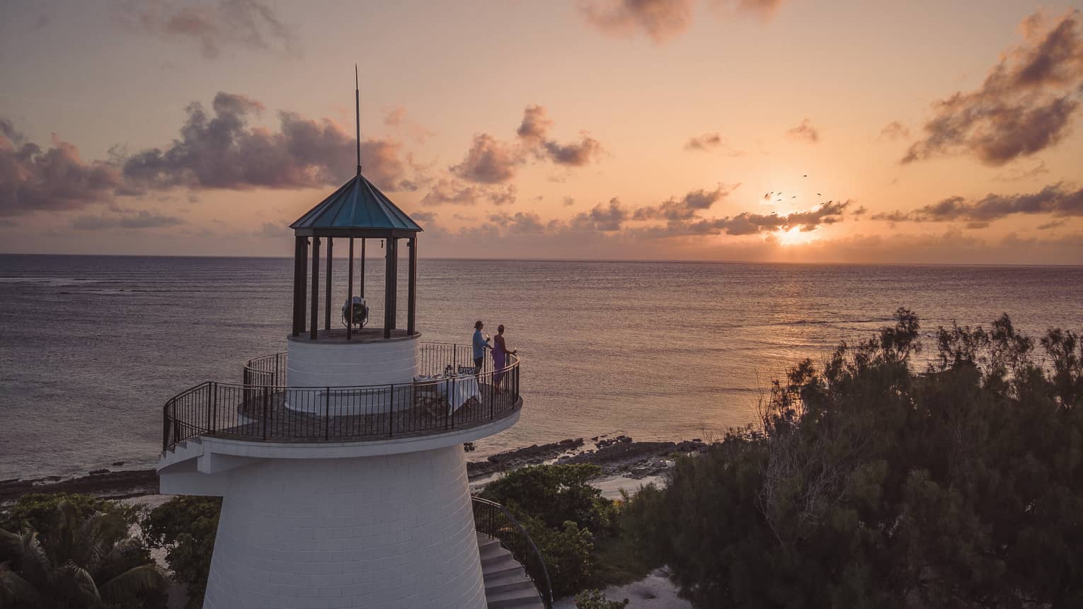 Long view of two people on a beach lighthouse balcony, enjoying the sunset over the ocean, distant silhouette of birds. 