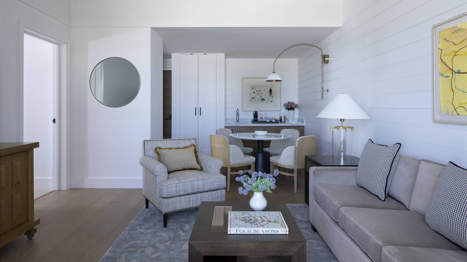 Guest room living area with tan sofa and armchair, ergonomic wooden coffee table, dining table for four, patterned area rug, doorway to bedroom