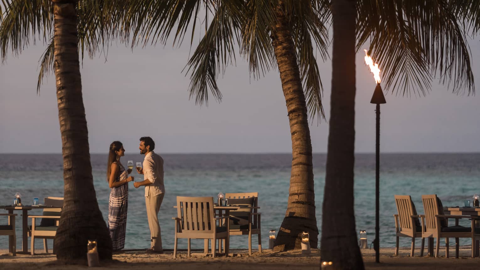 A couple talks on the beach at sunset, a tiki torch burns, table and chairs sit on beach