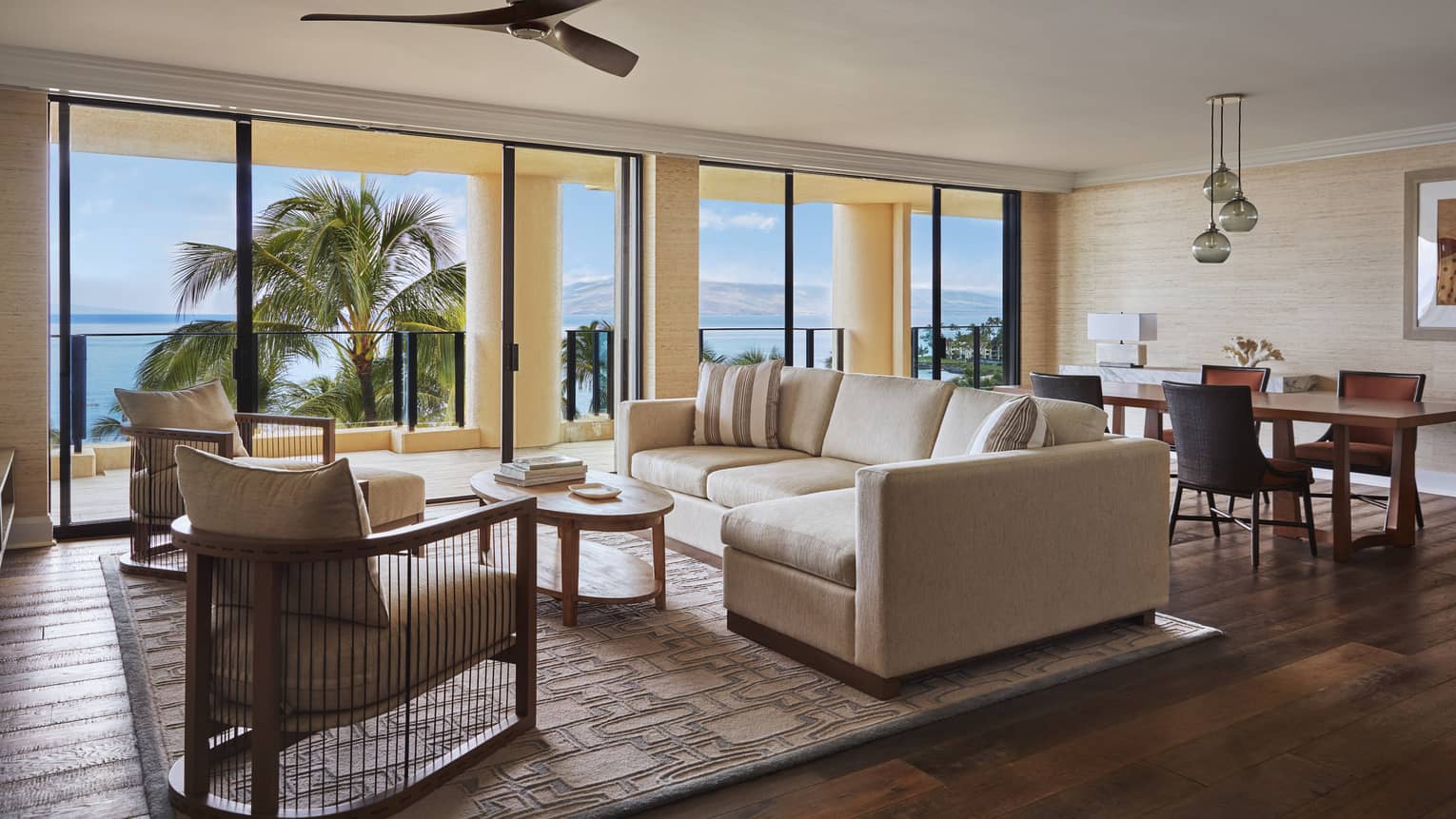 Spacious living and dining area with a beige sofa, armchairs and dining table, opening to a balcony with ocean and palm trees view at a luxury resort suite