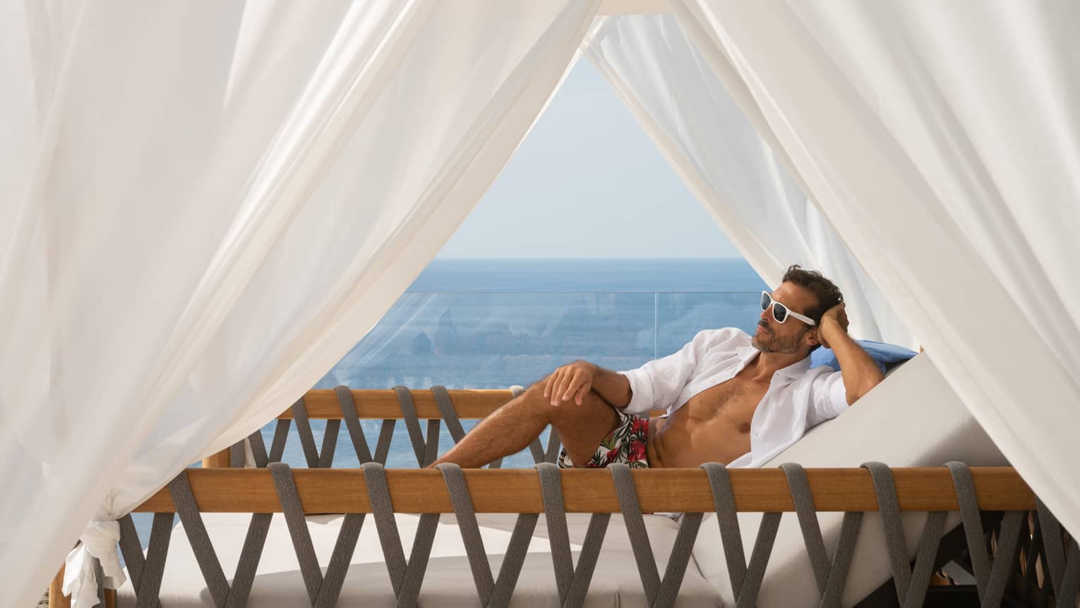 A person reclines on a lounge chair in a cabana with white curtains, with ocean views in the background.