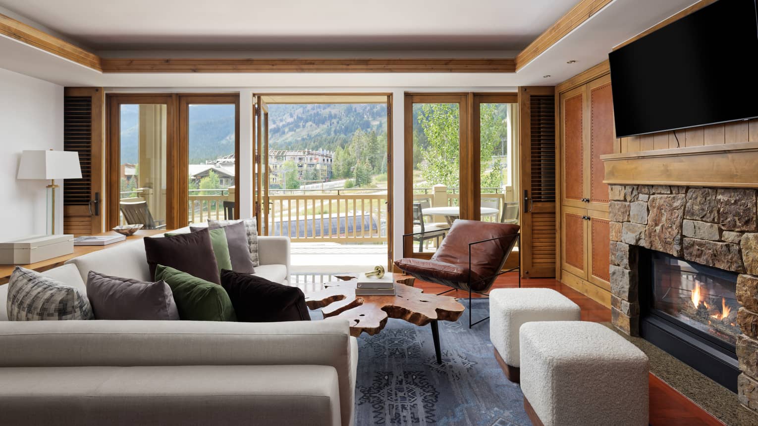 Luxury suite living area with a grey sectional sofa, rustic wood coffee table, stone fireplace, TV, leather chair, cream ottomans and walk-out balcony