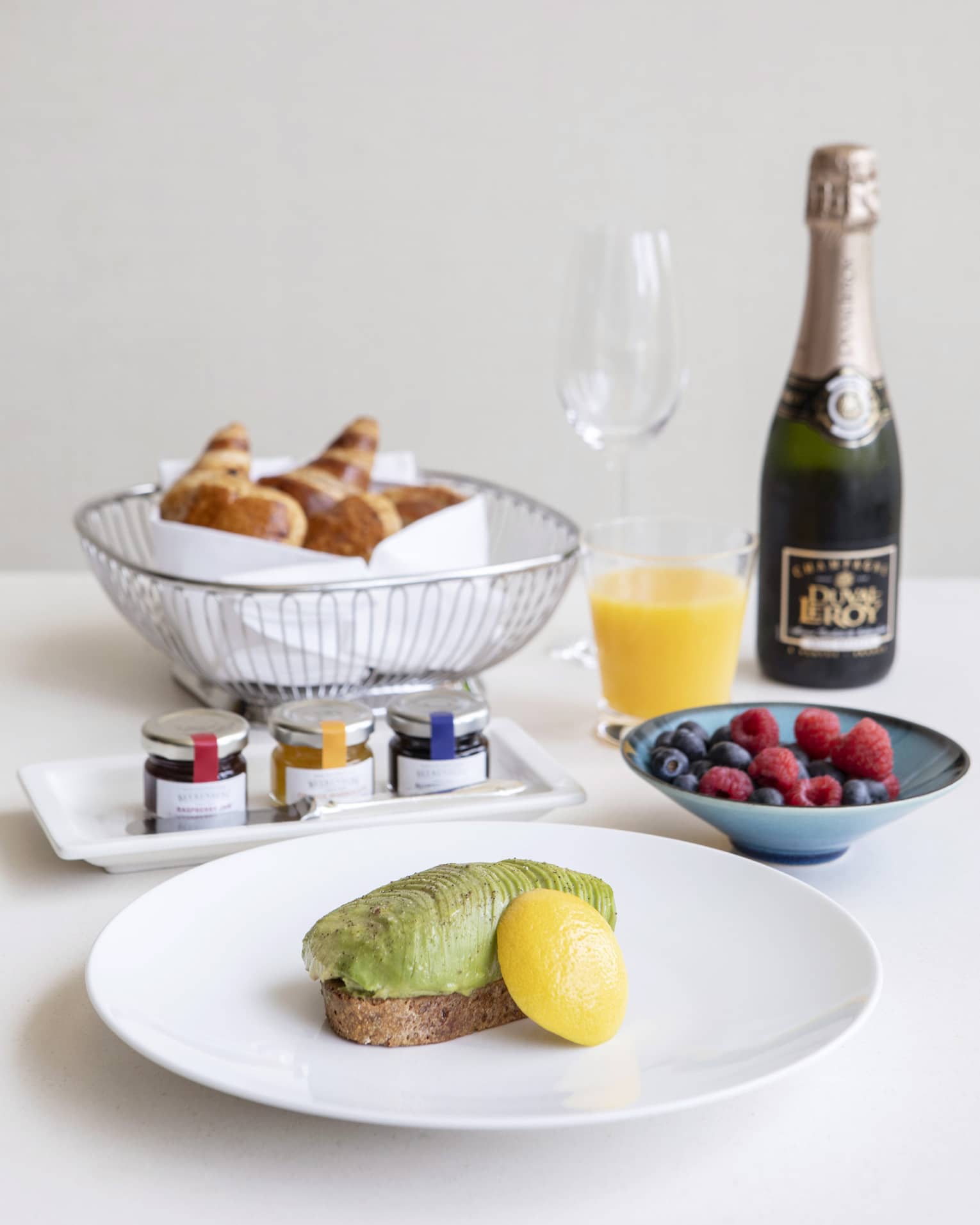 Luxurious breakfast including avocado toast, a basket of croissants, fresh fruit, and champagne and orange juice.