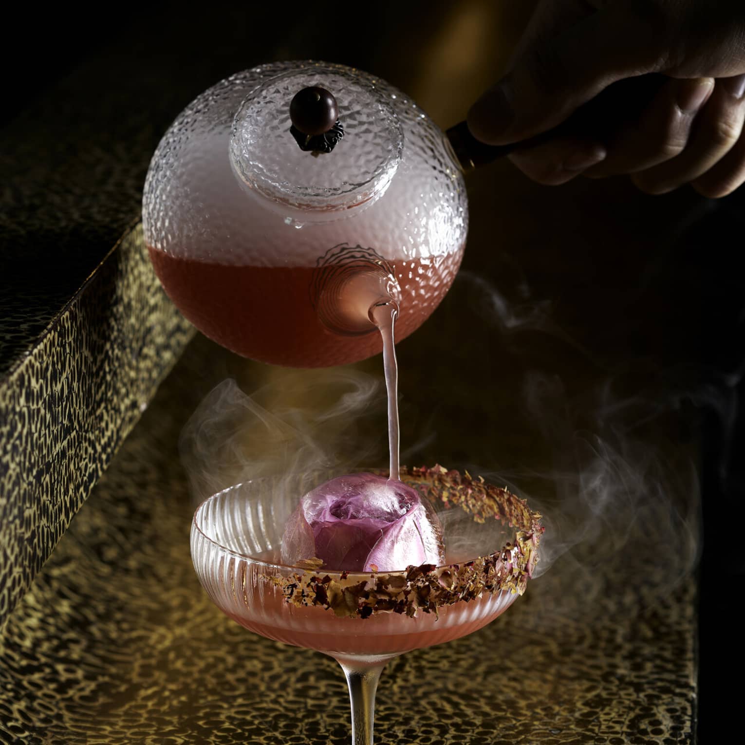 A vibrant pink cocktail being poured from a textured glass teapot into a coupe glass with a large ice sphere, garnished with a rim of crushed spices.