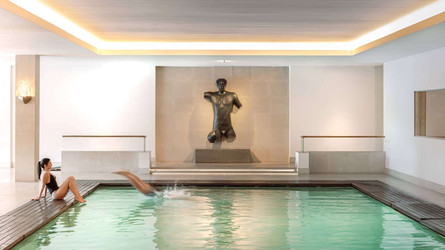 An indoor pool in a bright space finished in neutral tones. A figurative sculpture is displayed against a wall at the end of the pool.