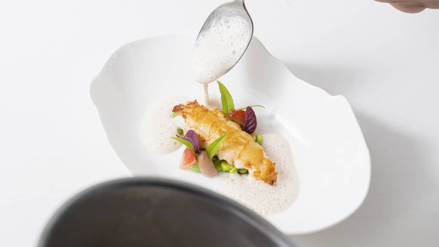 A spoon places foam around a piece of fish served on a bed of greens