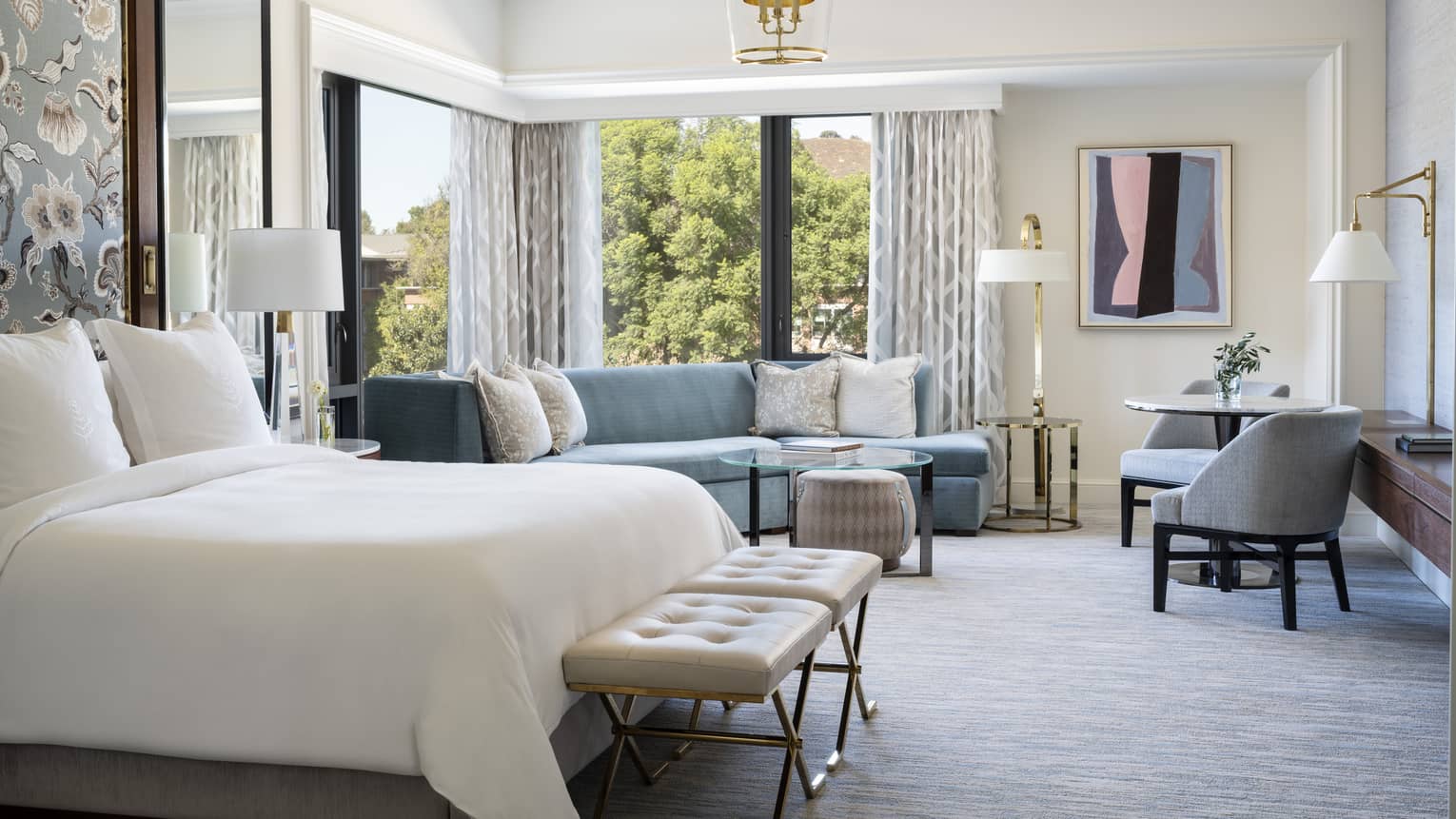 Sunlit corner studio suite with blue furnishings, carpet, drapes, view of trees.