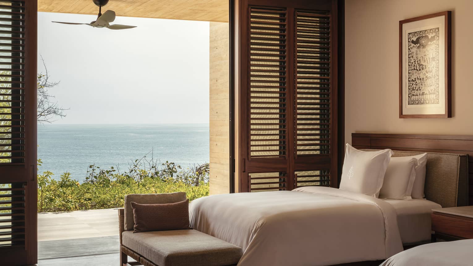 Bedroom with twin beds and white linens, opening to a terrace with ocean views. Wooden shutters and a ceiling fan enhance the room’s airy, tropical feel.