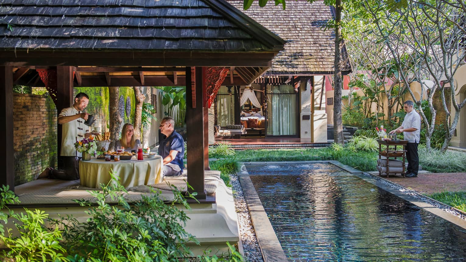 Hotel staff serve couple under dining gazebo by pond 