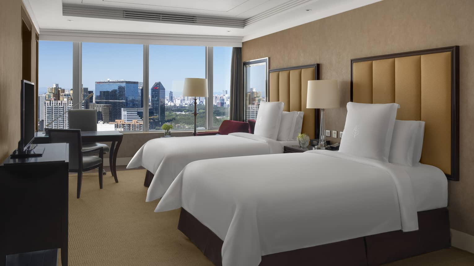Hotel room with two single beds, tan headboards, dresser and TV, desk and two chairs