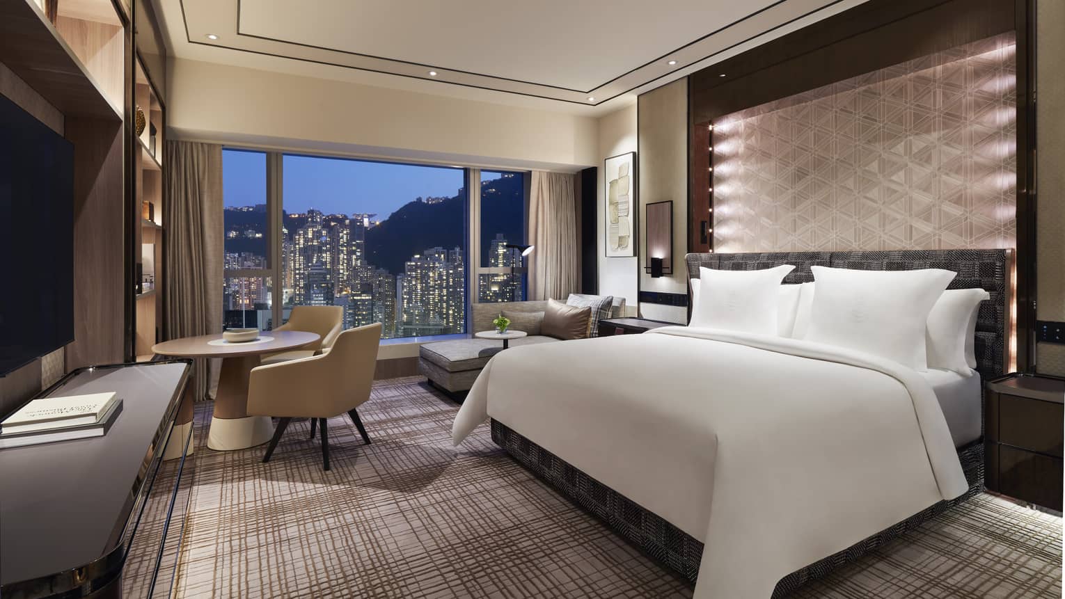 Luxury hotel room with a large bed, geometric accent wall, and a seating area. Floor-to-ceiling windows provide a nighttime view of city skyscrapers.