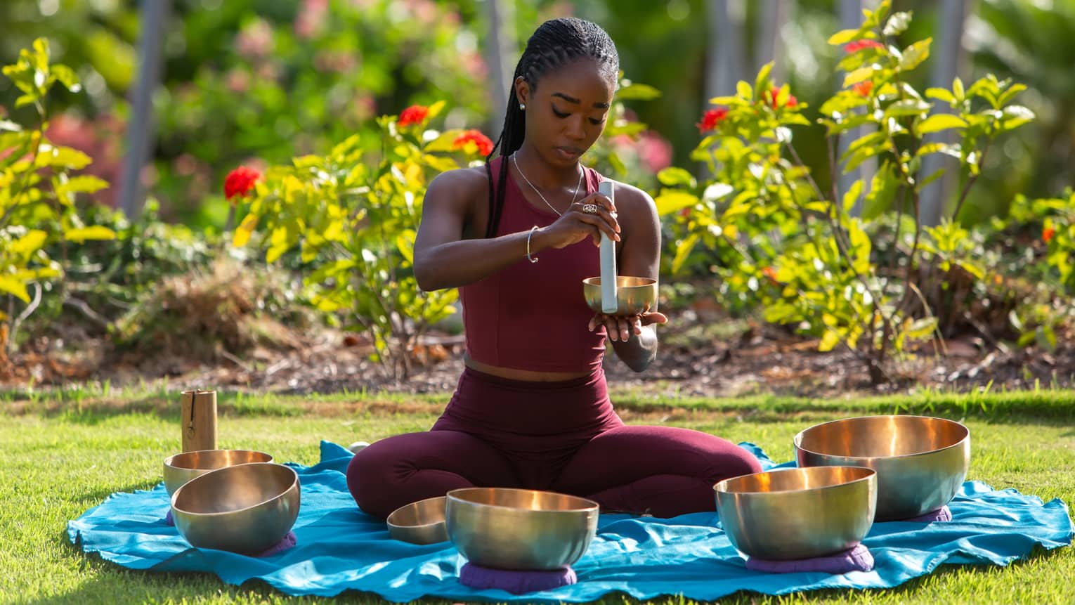 In a sunlit garden, a sound-bath practitioner sitting on a mat in a circle of brass bowls holds a tube against a small bowl.