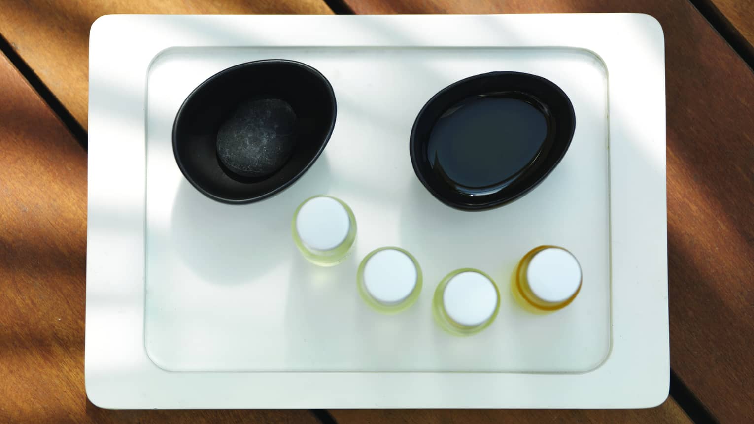 Aerial view of white spa tray with two black bowls with water, four small jars of oil