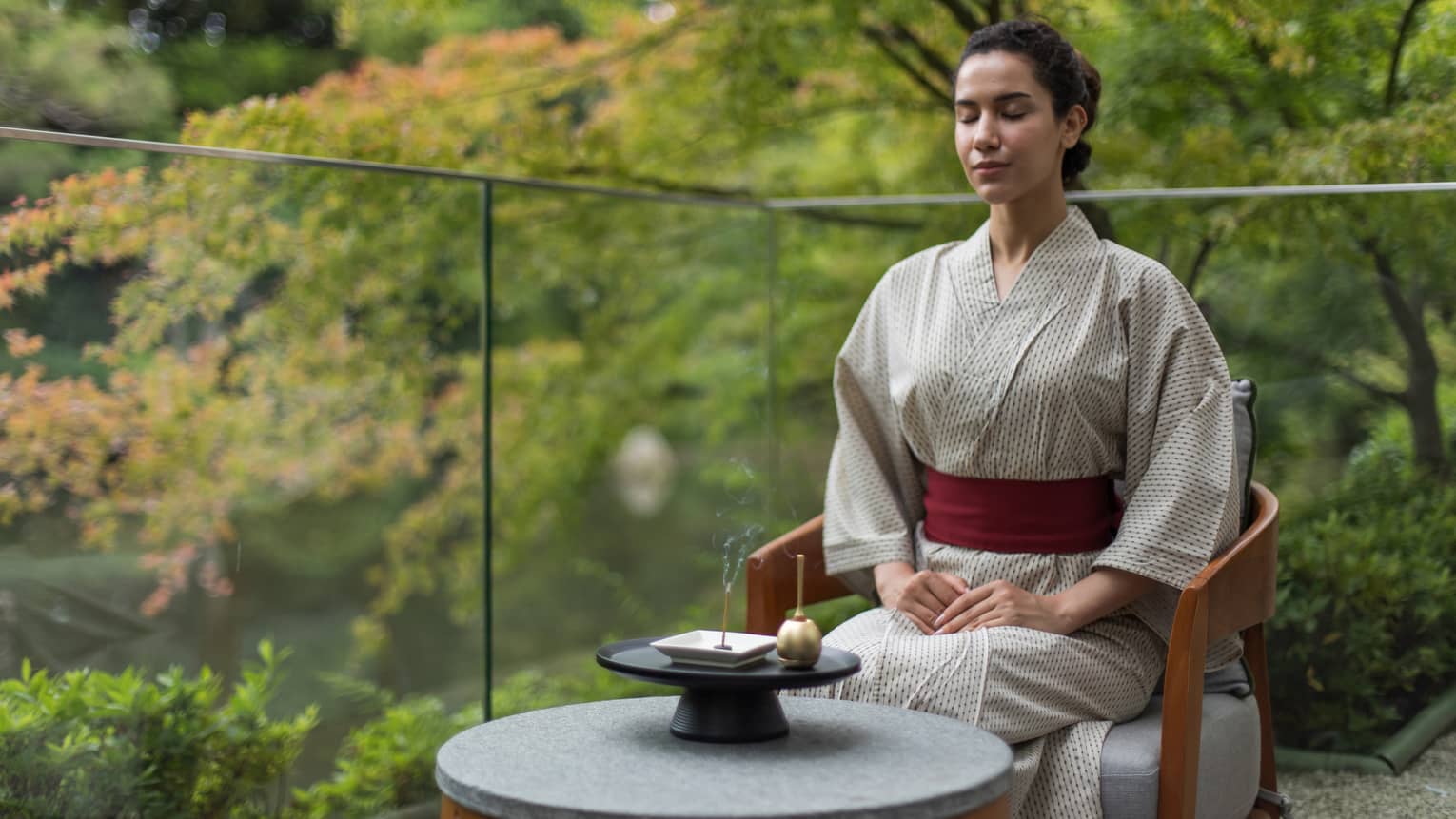 A woman sits on glass-enclosed balcony in lush setting with traditional Japanese robe on, meditating with an orin bell
