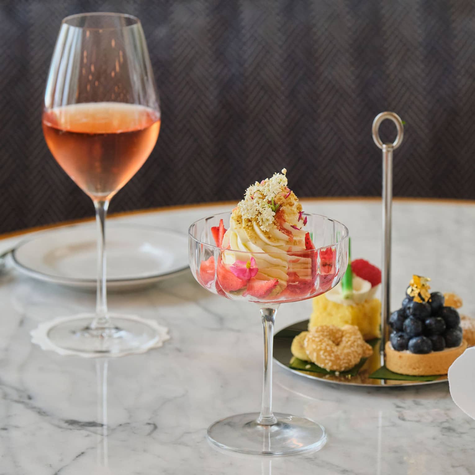 Ice cream served in a coupe glass sits next to a glass of wine and a small platter of desserts, all on a marble table