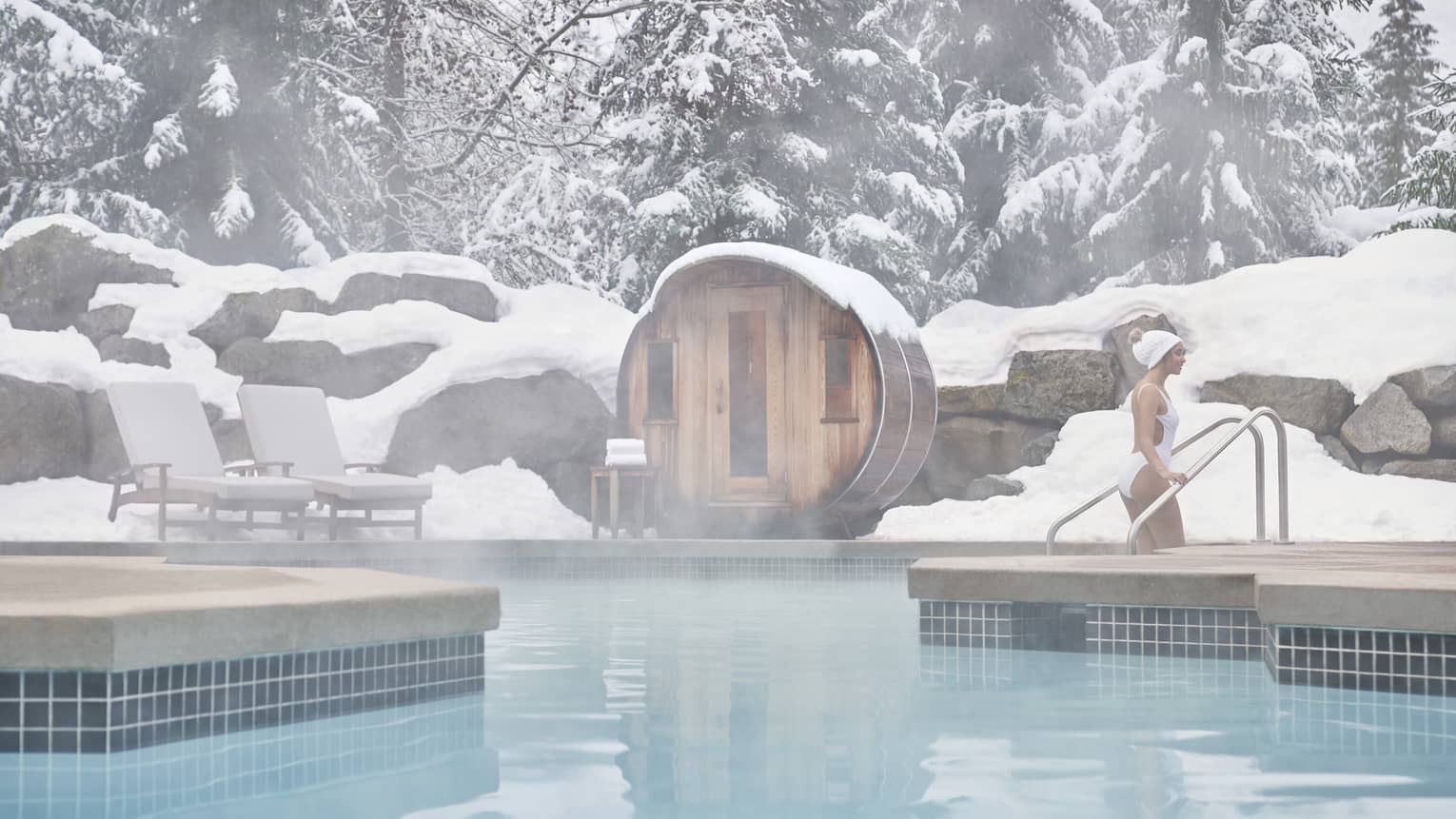 Woman in a white bathing suite and white hat steps out of an outdoor pool, surrounded by snow-covered boulders and trees in the background, with a circular sauna set right next to the pool