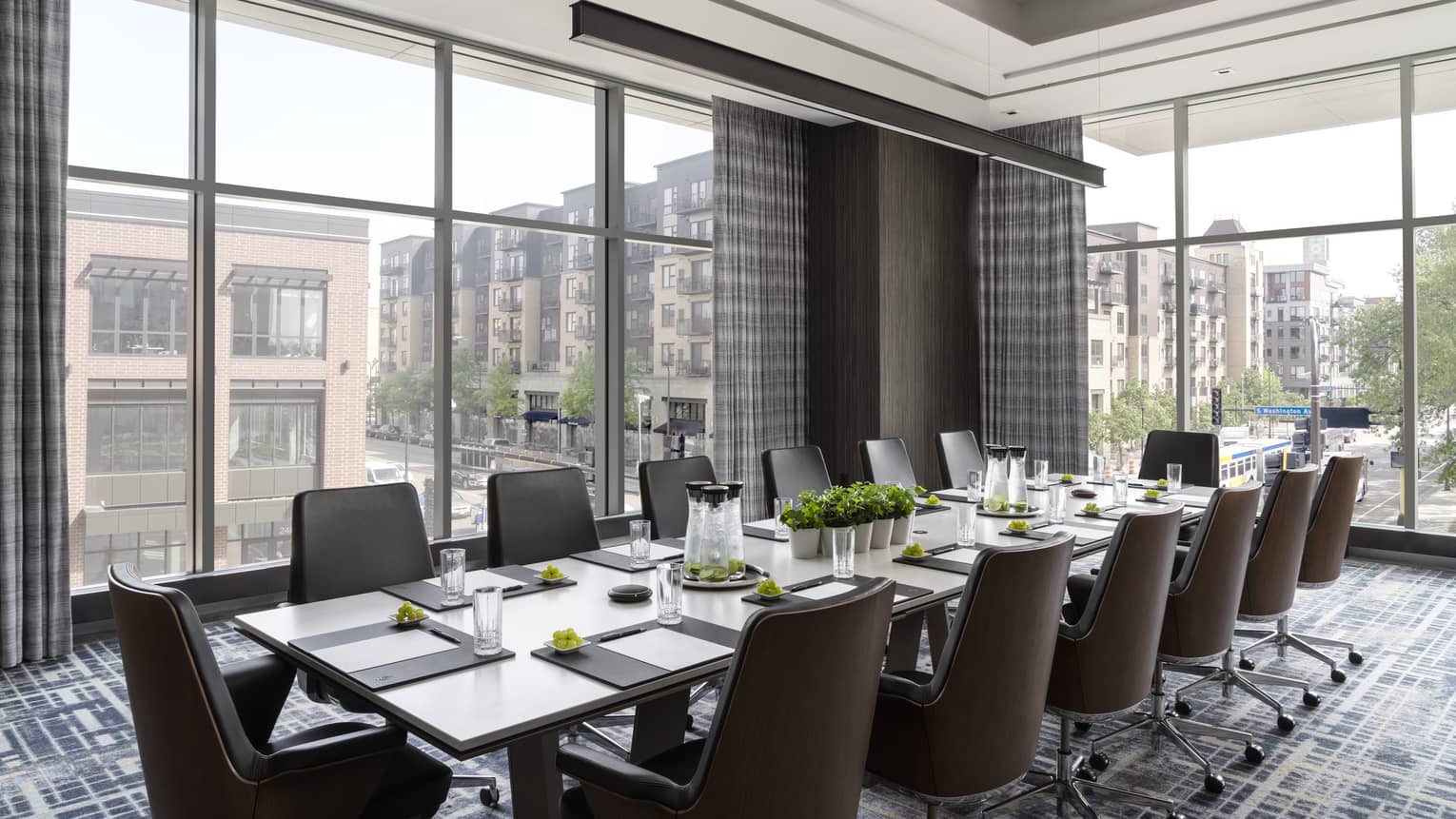 A meeting room with large windows, a long table, brown chairs and papers and supplies on table.