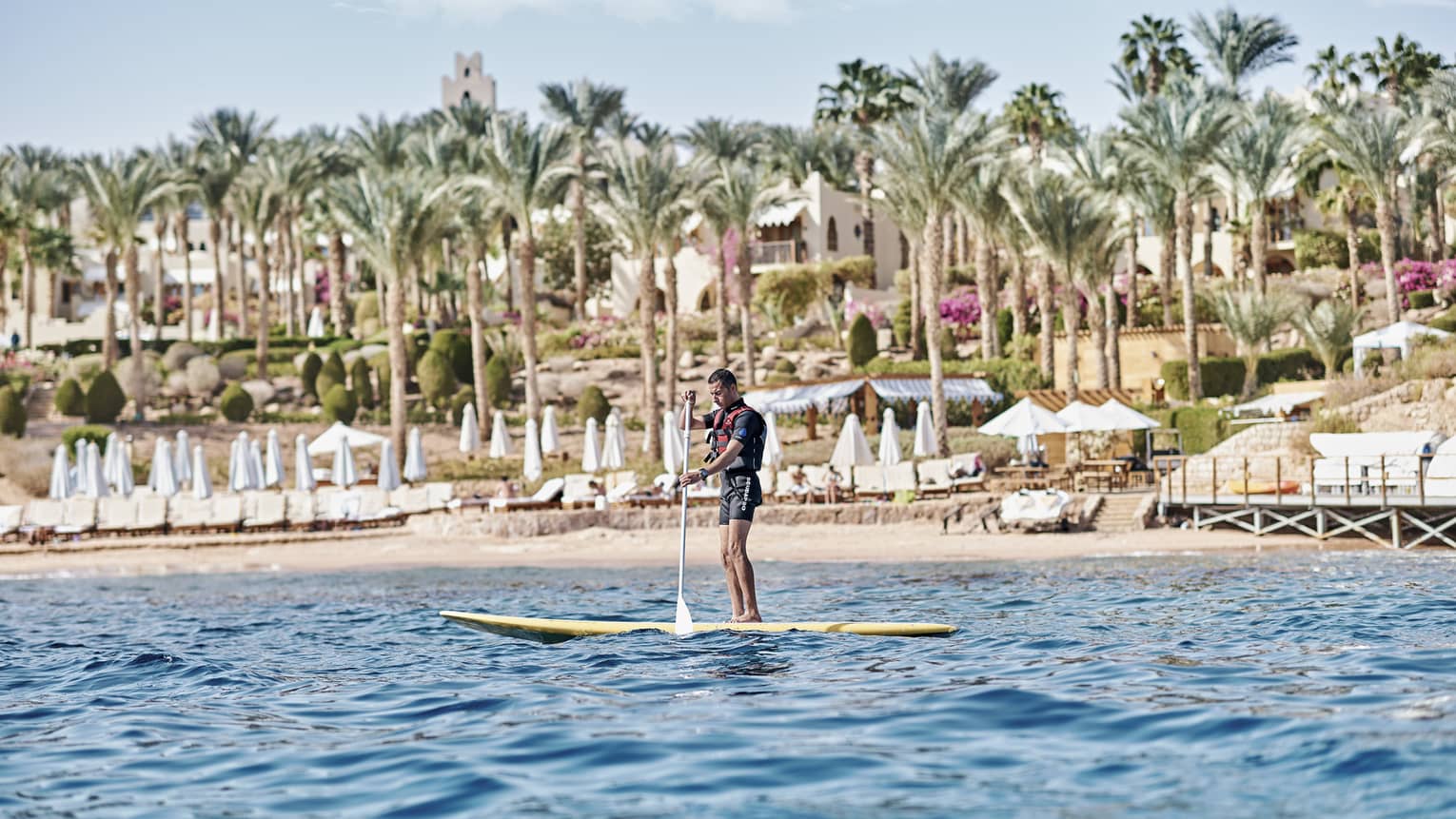 A paddler stands on a board in the water in front of a beach lined with lounge chairs and palm trees and a villa beyond.