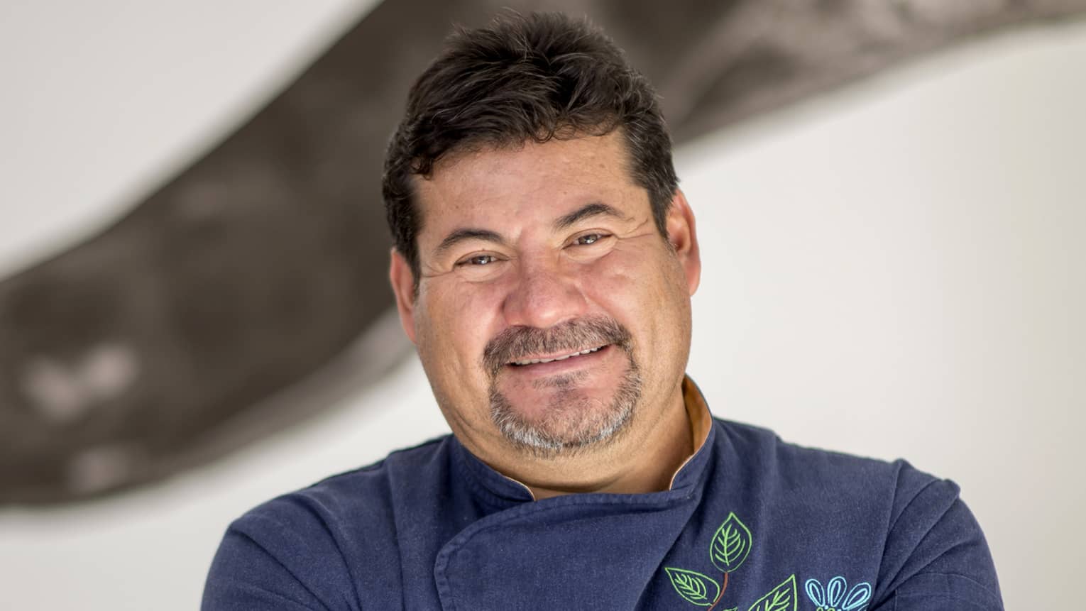 A chef wearing a navy embroidered coat with their arms crossed and smiling