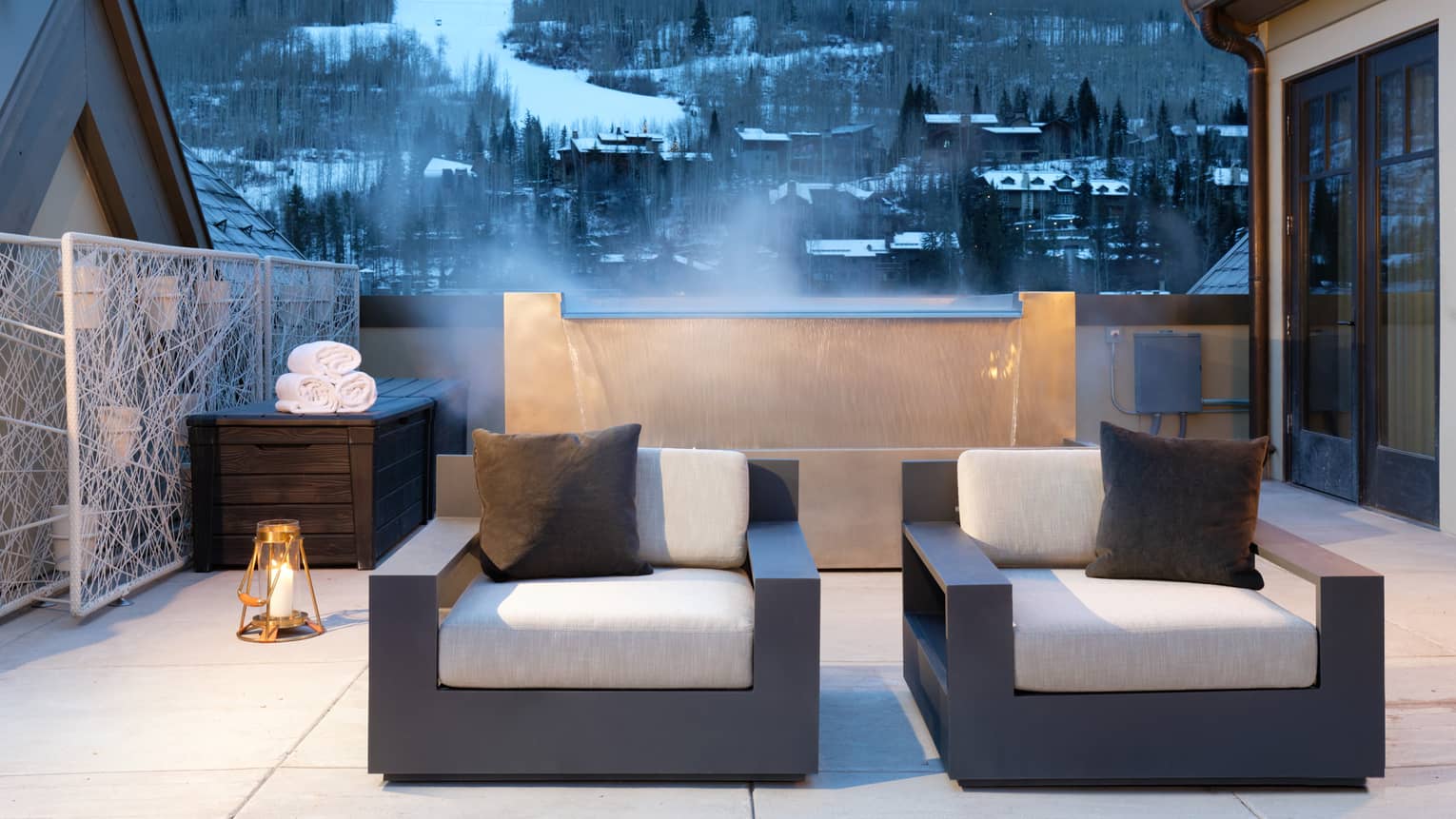 Plush patio armchairs in front of steaming fountain, snowy mountains in background