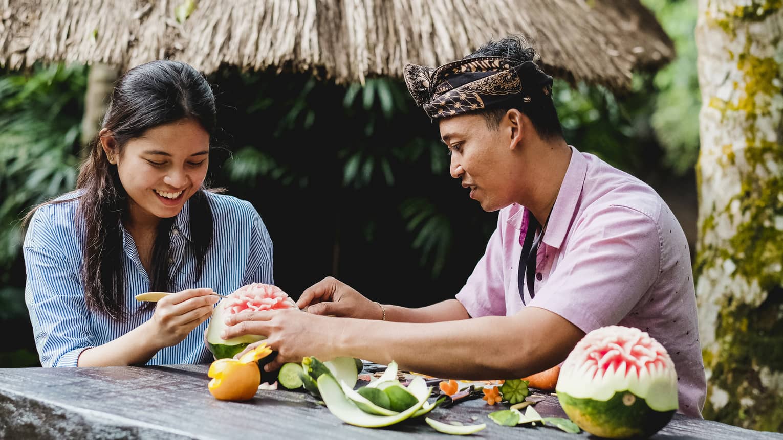 Two people carve a watermelon; beside them is a completed melon, its rind pared back to create an elaborate flower.