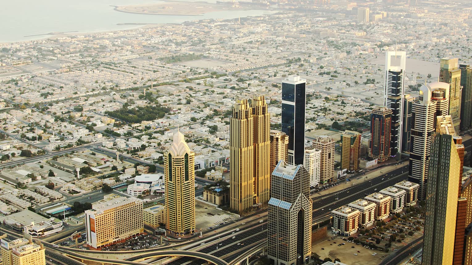 Aerial view of rows of golden towers reflecting sun against backdrop of low city grid receding into harbour under hazy sun.