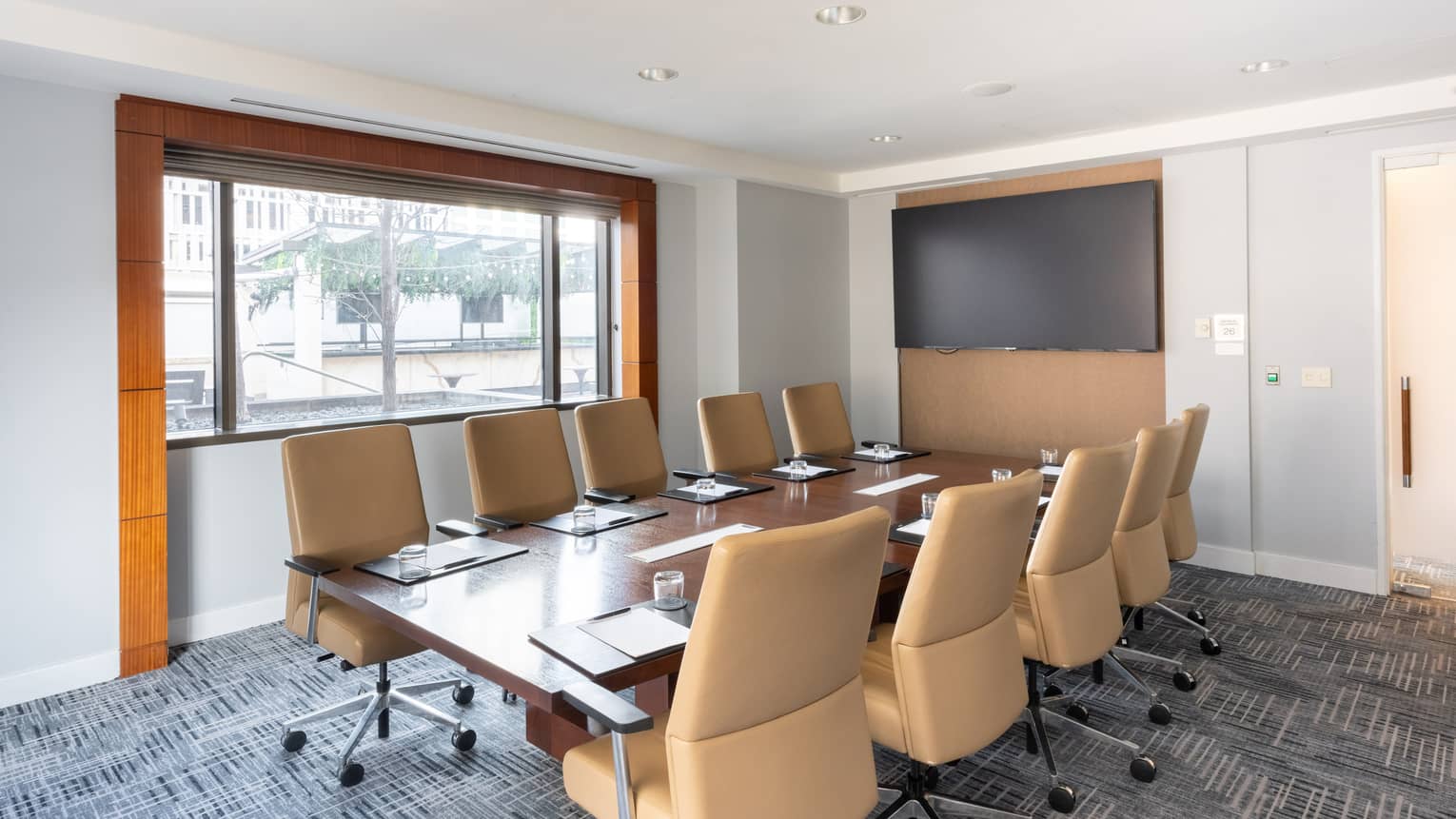 A serene meeting room with neutral decor, a long table, upholstered swivel chairs, recessed lighting in the ceiling, a large monitor on one wall, and a large window letting in natural light