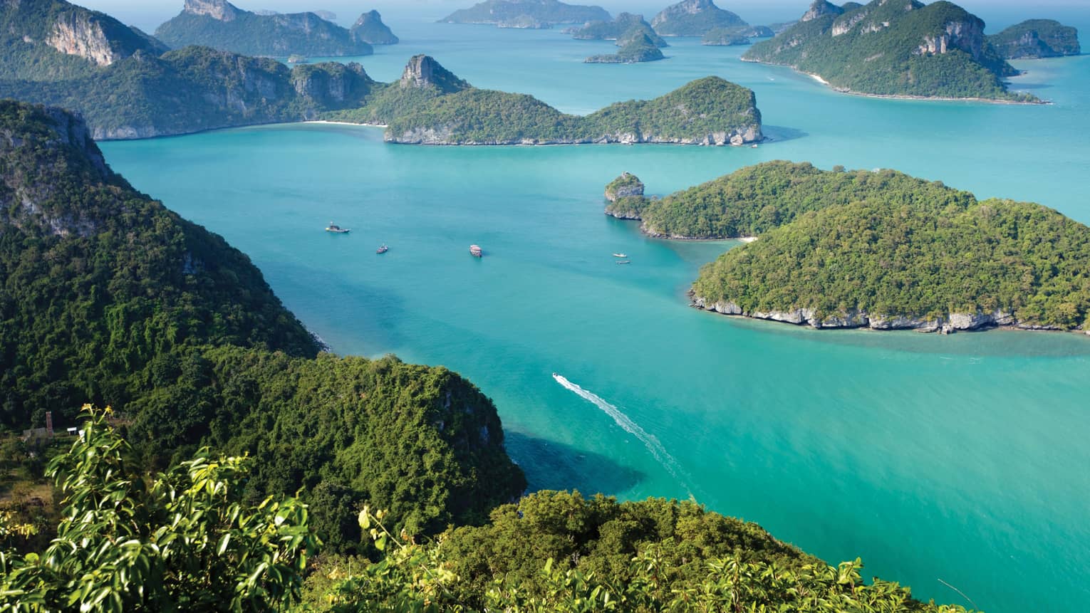 Aerial view of a dozen lush, forested, hilly islands of different sizes surrounded by calm turquoise water dotted with boats.
