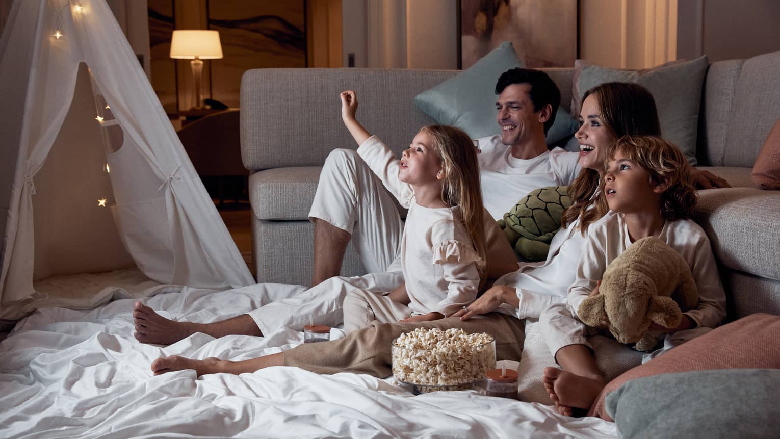 A family relaxing on blankets beside a sofa; the children hold stuffed toys and there's a bowl of popcorn between them.