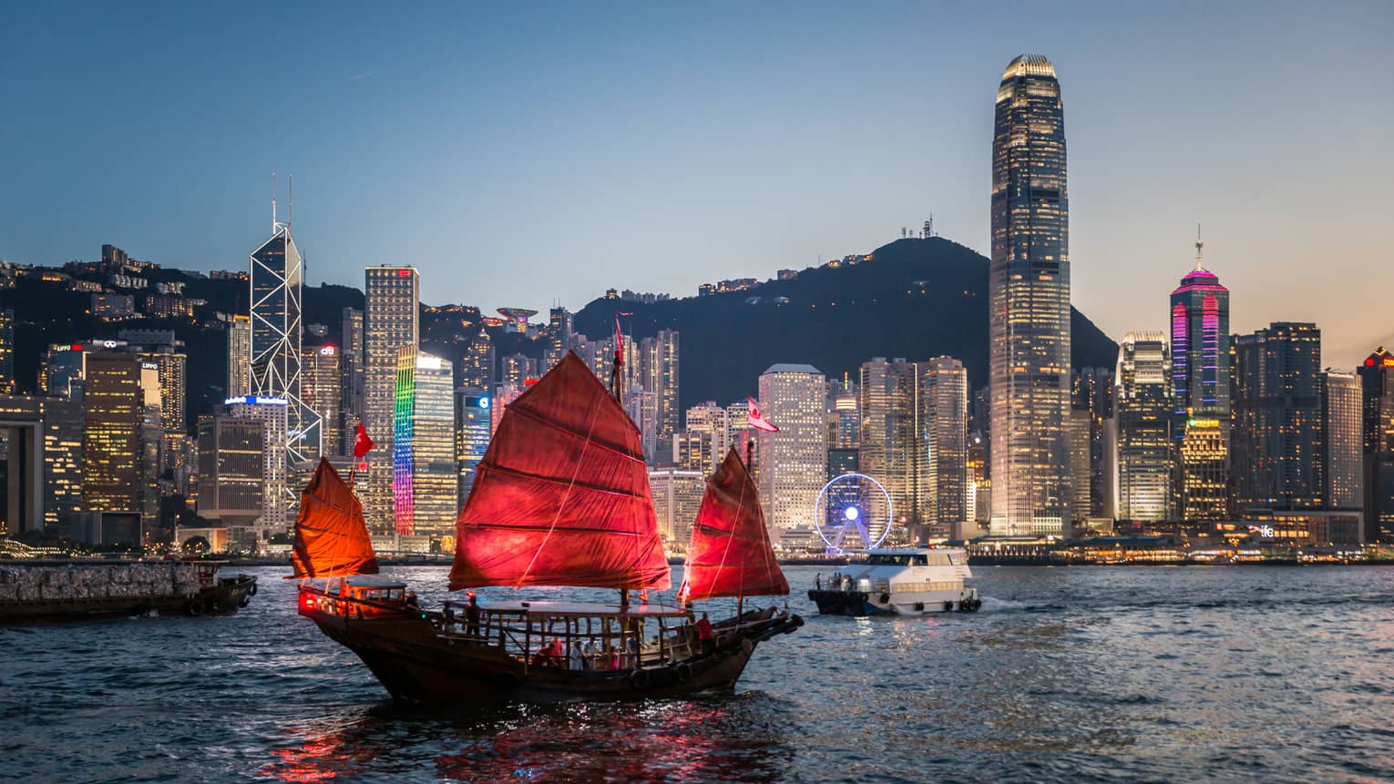 A traditional Hong Kong junk boat with red sails gliding through Victoria Harbour at dusk, with the city’s illuminated skyline in the background