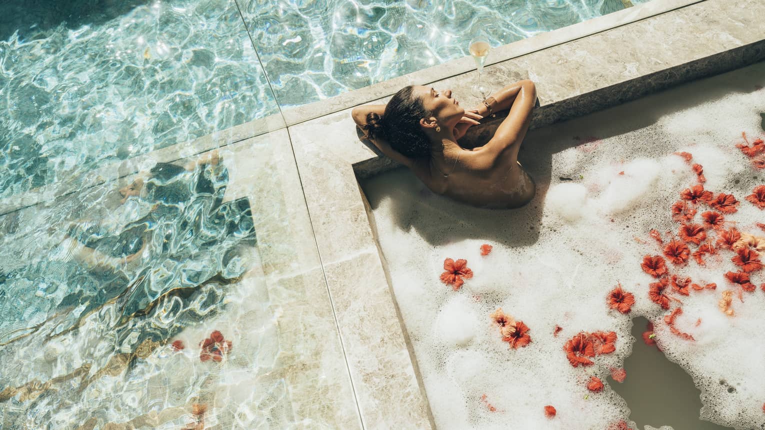 Surrounded by mirrored walls, a bather relaxes, eyes closed, in a marble tub of frothy water and red hibiscus blossoms.
