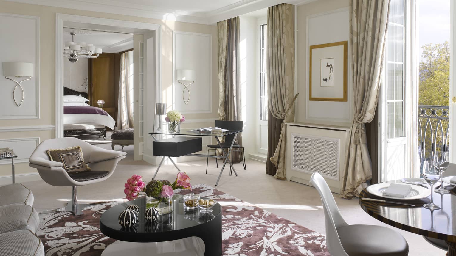 Luxury hotel suite living area with a modern armchair, black desk, round coffee table, dining table and balcony access, leading to a bedroom with a king bed