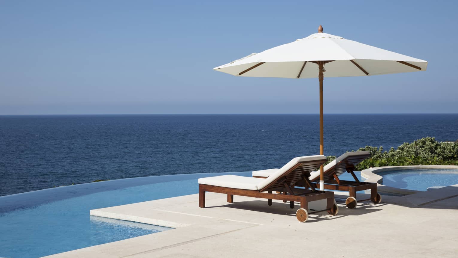 Two lounge chairs under white patio umbrella at edge of infinity pool, ocean views
