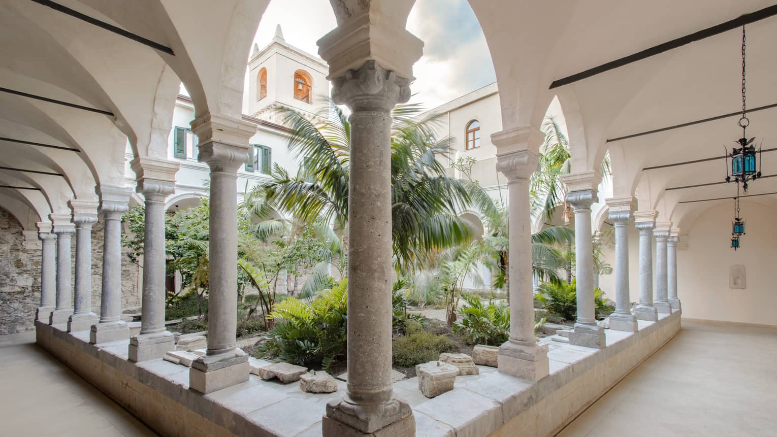Cloistered courtyard with stone columns, arches and lush greenery, framed by the architecture of a historic building.