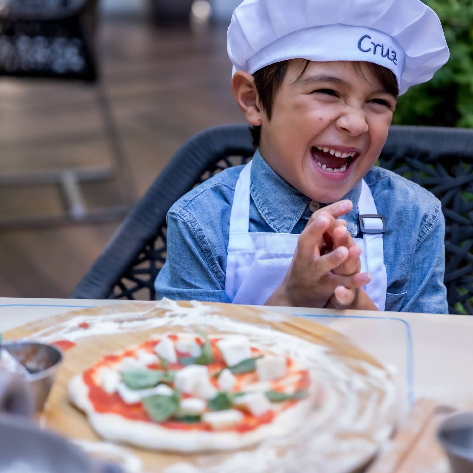 A young boy in a chef's hat laughing while making a pizza.