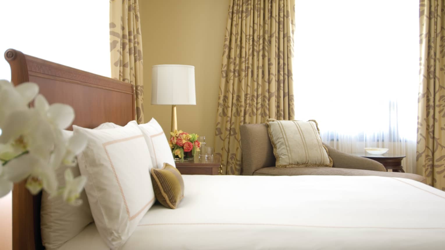 Deluxe City-View Room white orchids, side of bed, pillows against headboard, armchair, window in background