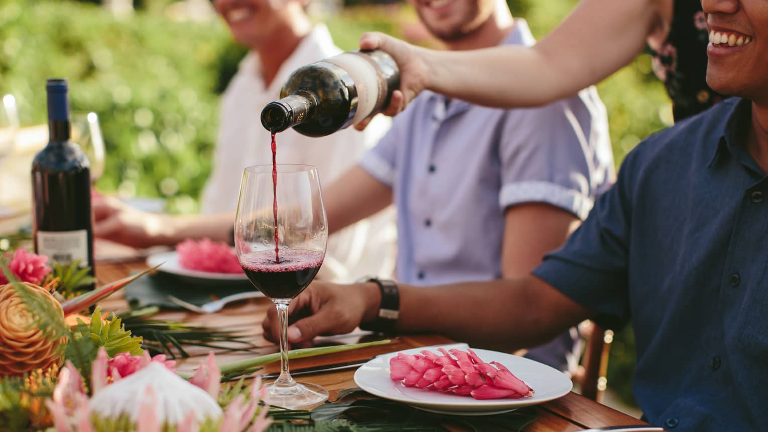 Three men sit at outdoor wooden table with plates of food, as server pours red wine into glass 