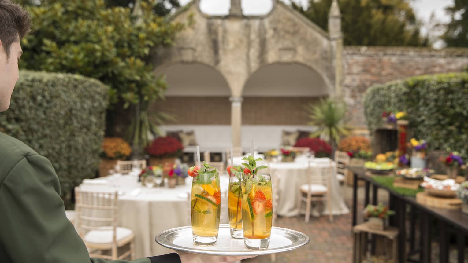 Hotel waitstaff holds tray with fruit-filled cocktails in garden event space 
