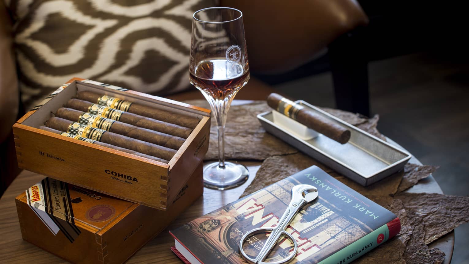 Box of cigars, book and glass of bourbon sit on small round wooden table