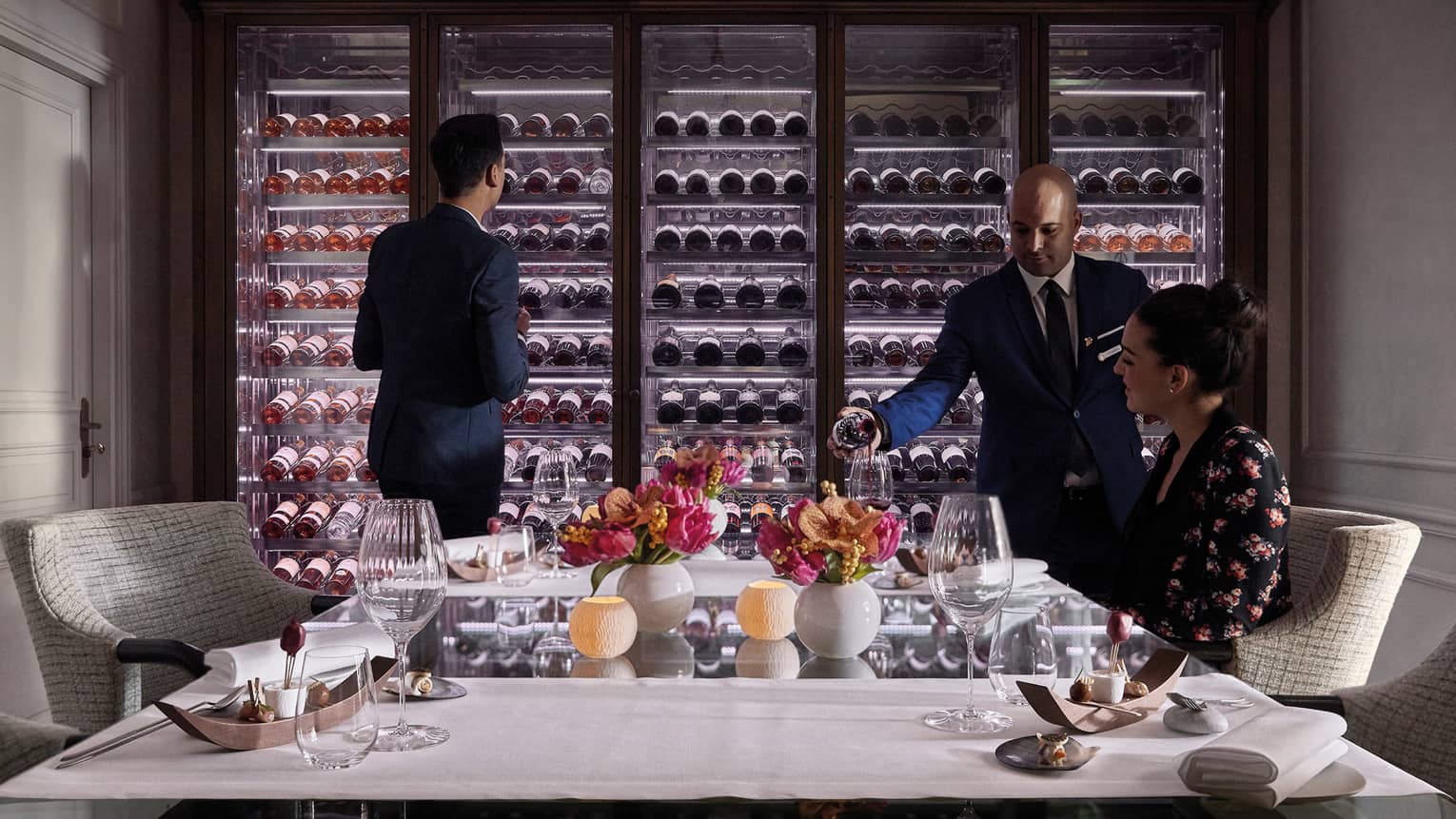 Man scans glass cellar as waitstaff pours wine in glass for woman at table