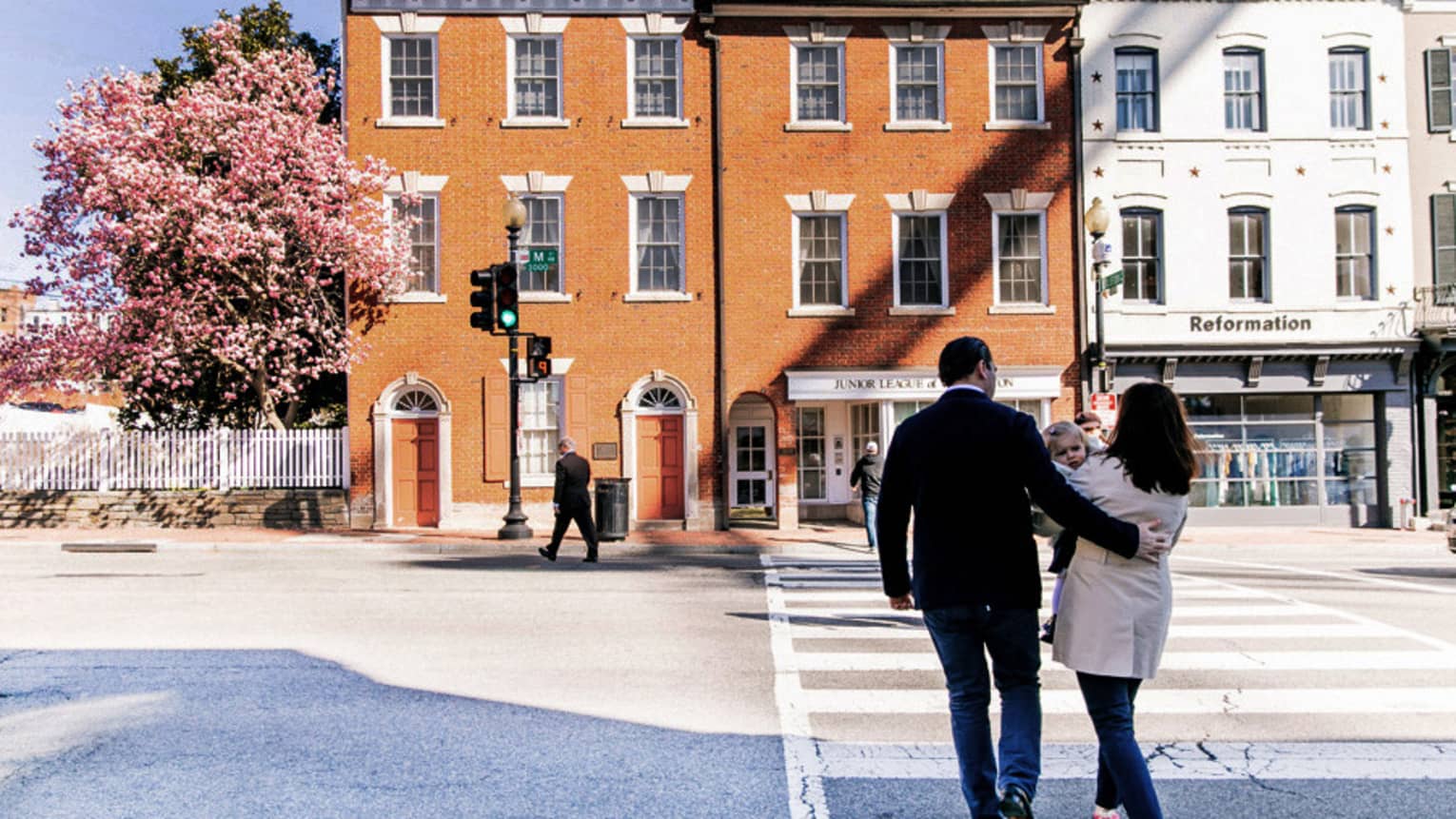 A man and woman walking along a cross walk towards an orange building with a cherry blossom tree next to it.