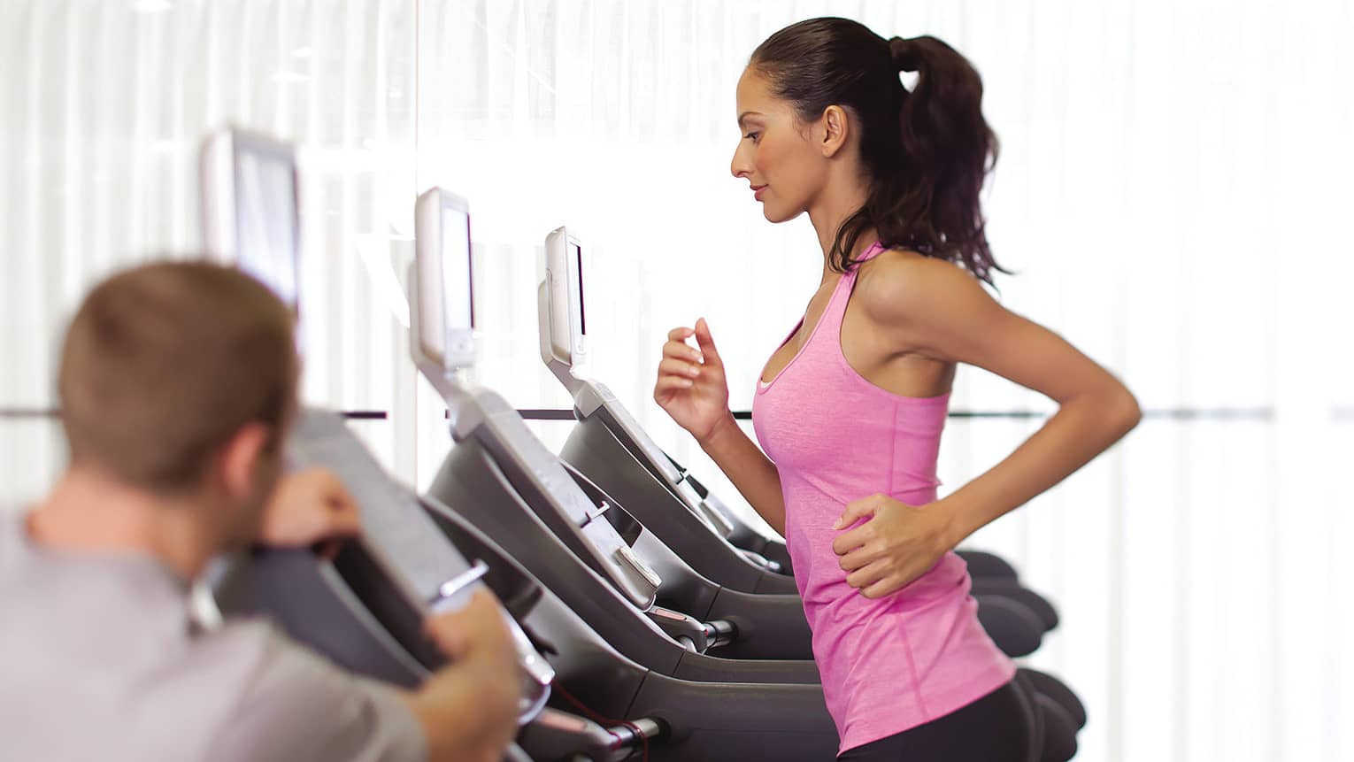 Woman in pink sleeveless shirt and ponytail runs on treadmill as personal trainer watches