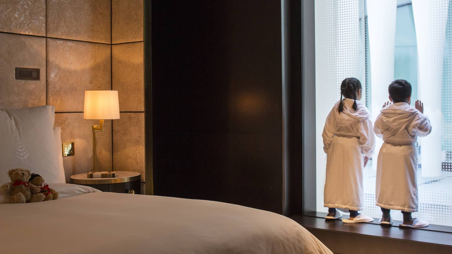 Young boy and girl in bath robes and slippers peer out the window at the city below, two teddy bears on the bed behind them