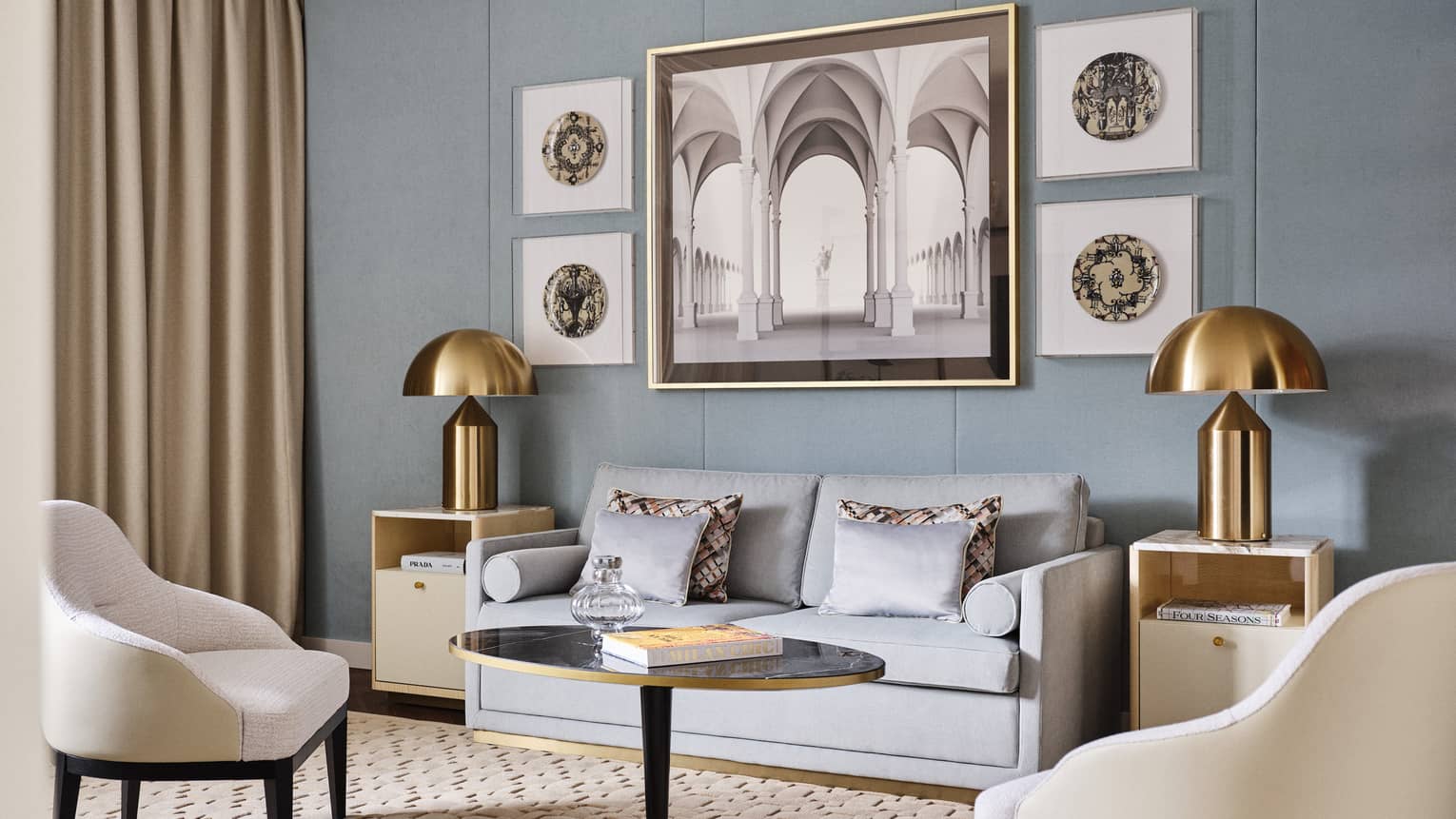 Contemporary living room of a hotel suite, featuring grey sofa, brass lamps, light blue wall with artwork