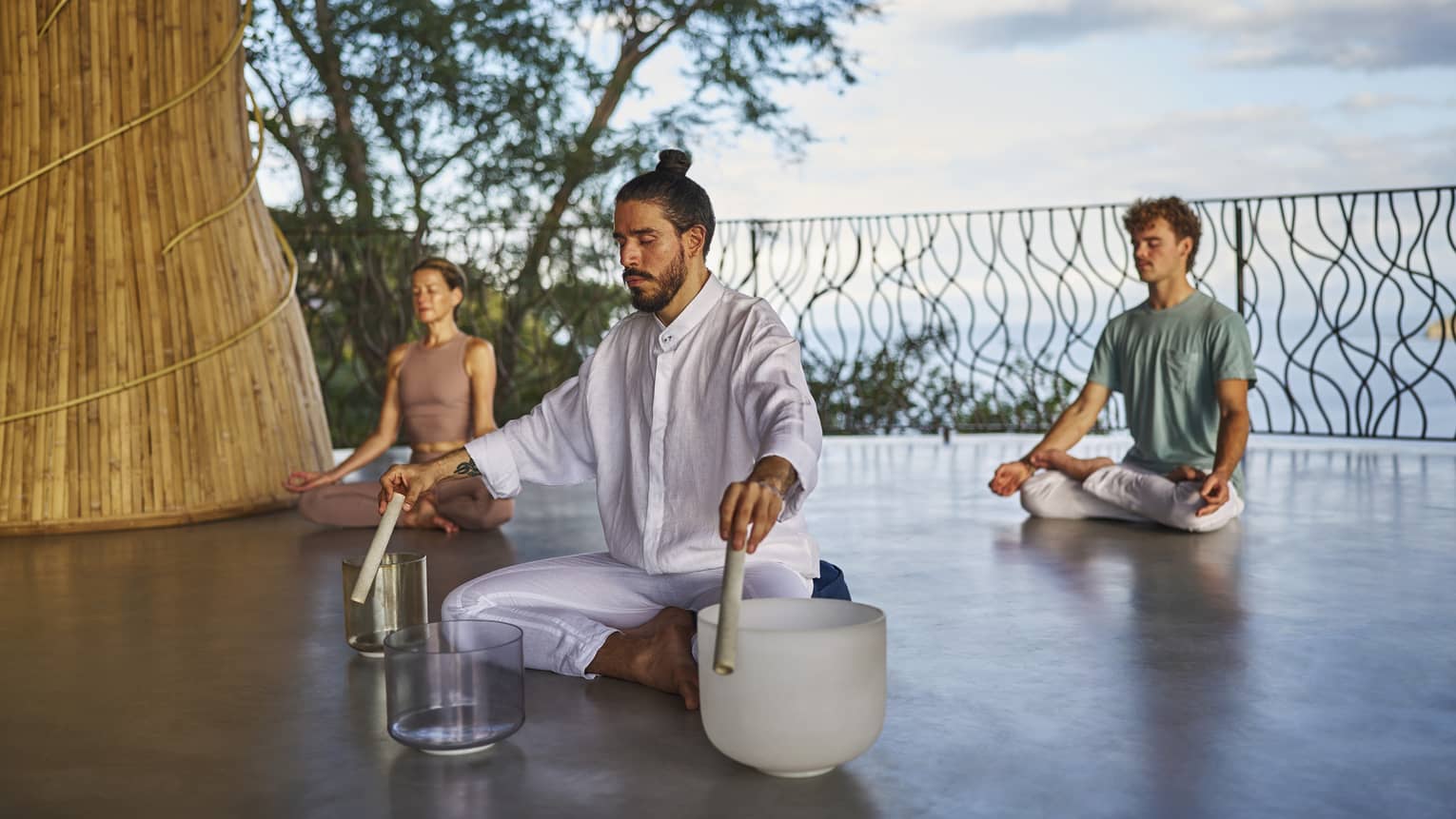Man wearing all white clothing sits cross-legged on floor with three singing bowls in front of him and two people also sitting cross-legged on the floor behind him