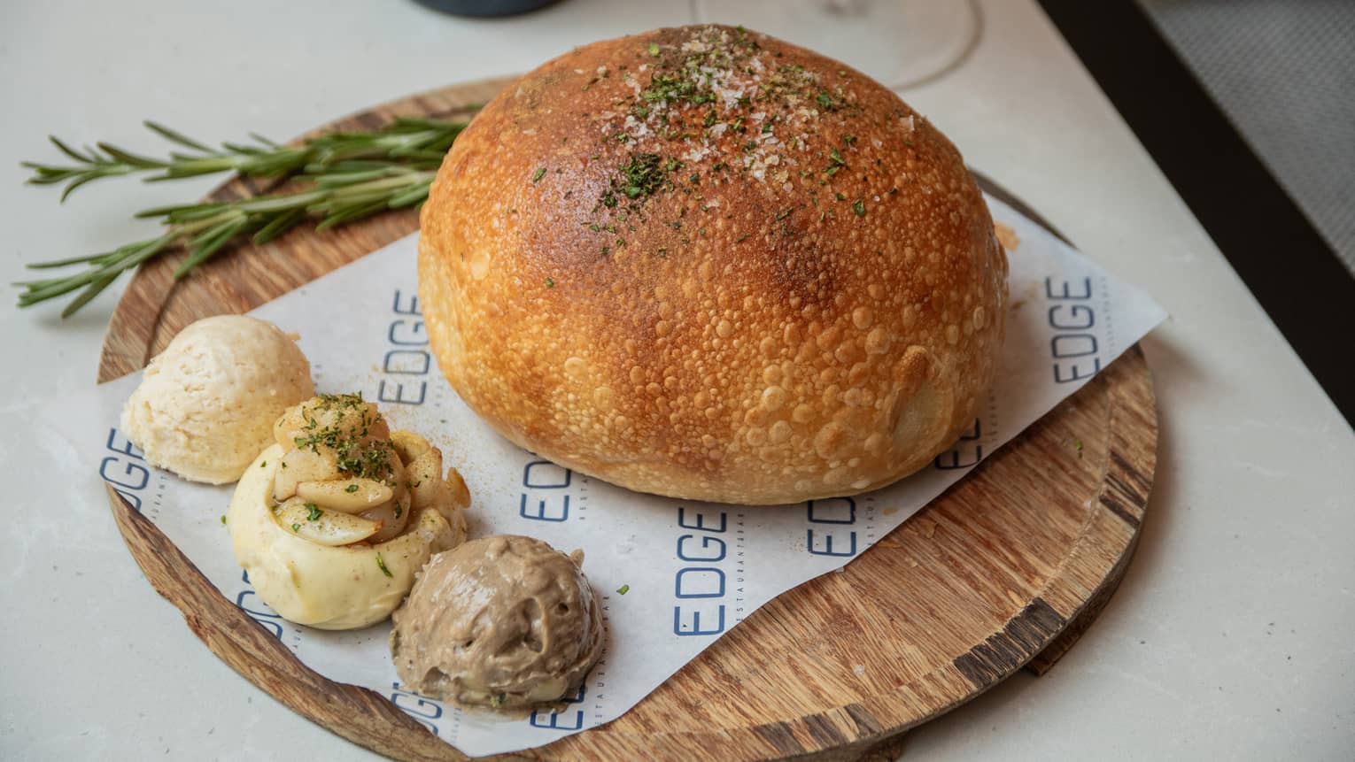 Round loaf of bread and three scoops of different kinds of spreads are plated on a round wooden platter along with a sprig of greenery