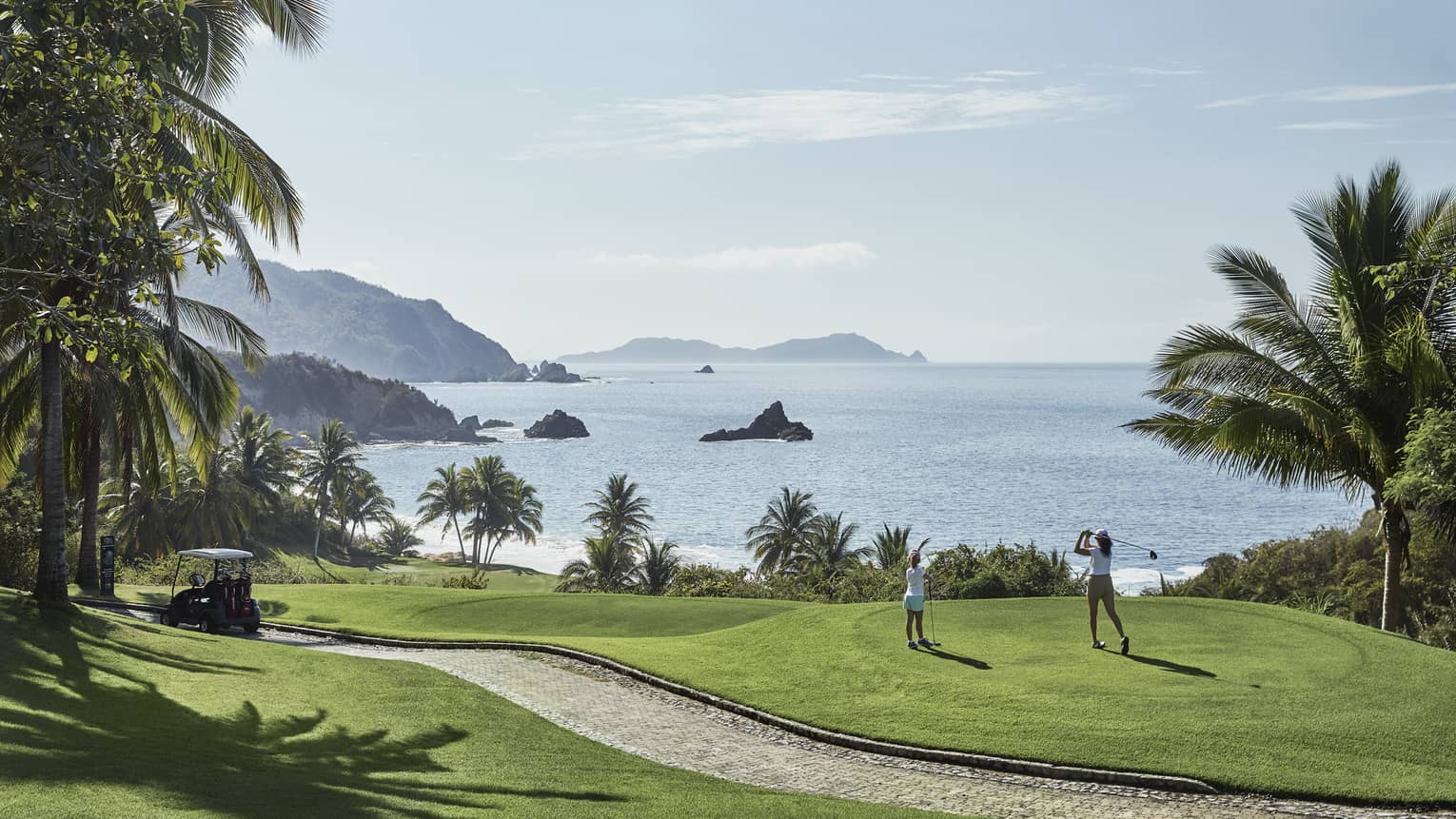 A guest swings from the tees on a hilly fairway bordered by palm trees and blue water with rocky outcrops as a child watches.