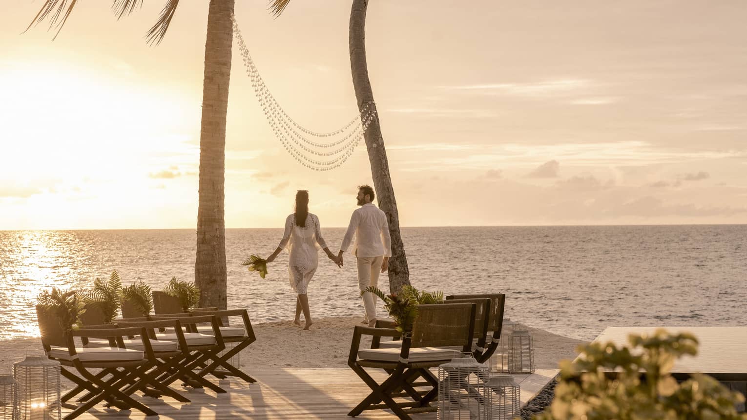 Couple wearing white hold hands under two palm trees with lights, looking at ocean