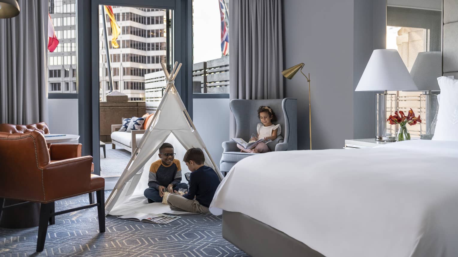 Children sitting in a tent and on a chair in a hotel room.